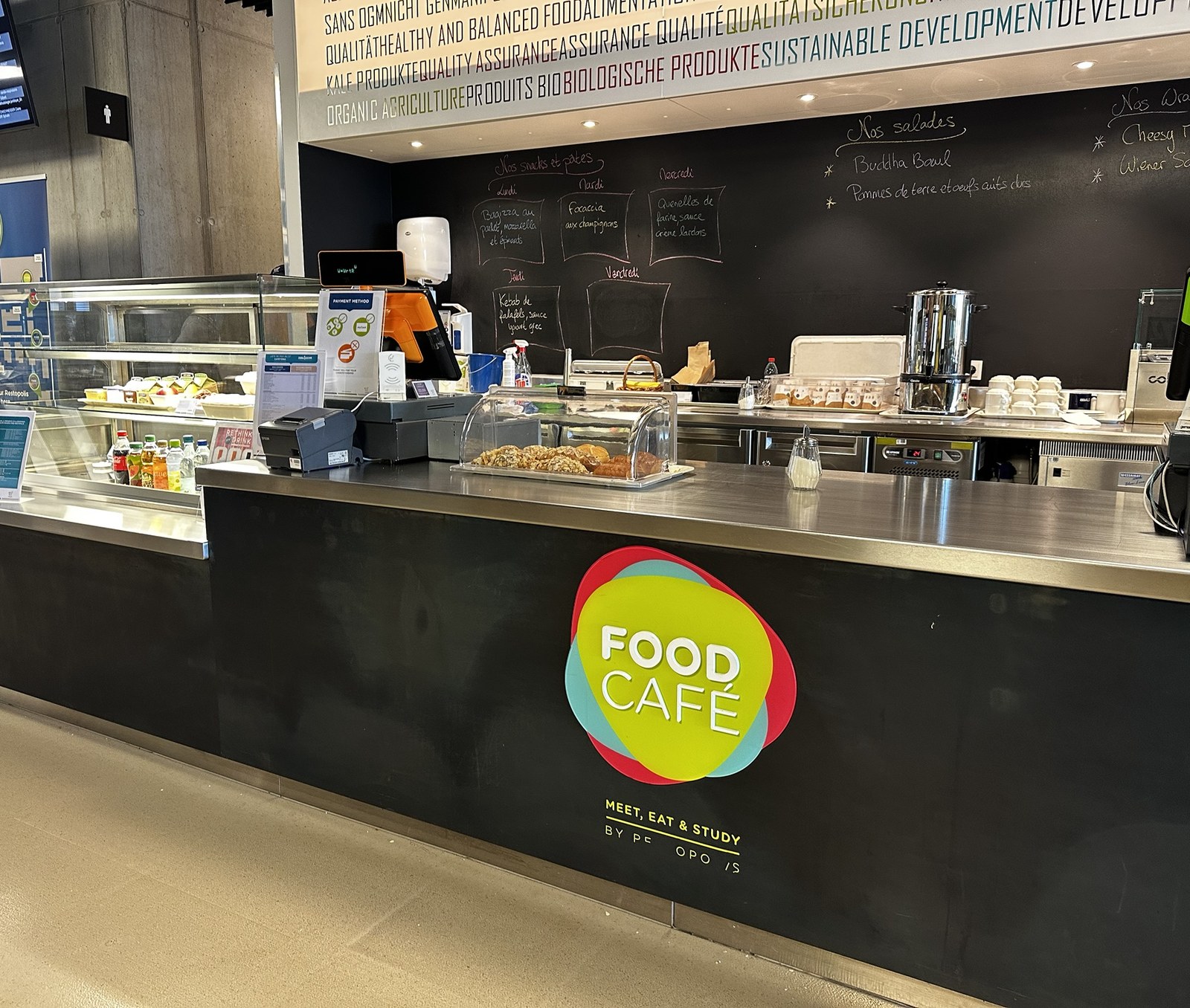 The Food Café counter on the Belval campus, its yellow and red roundel on the front, pastries and drinks in the chiller and a chalkboard of the week’s snacks behind.