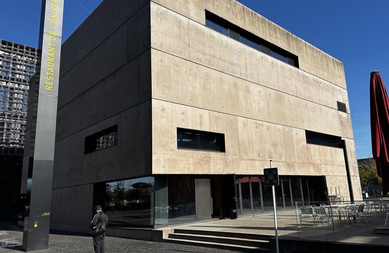 The Restaurant de l’Université building on the Belval campus, a concrete block with its name on a tall totem and a terrace of tables under red parasols beside the water.