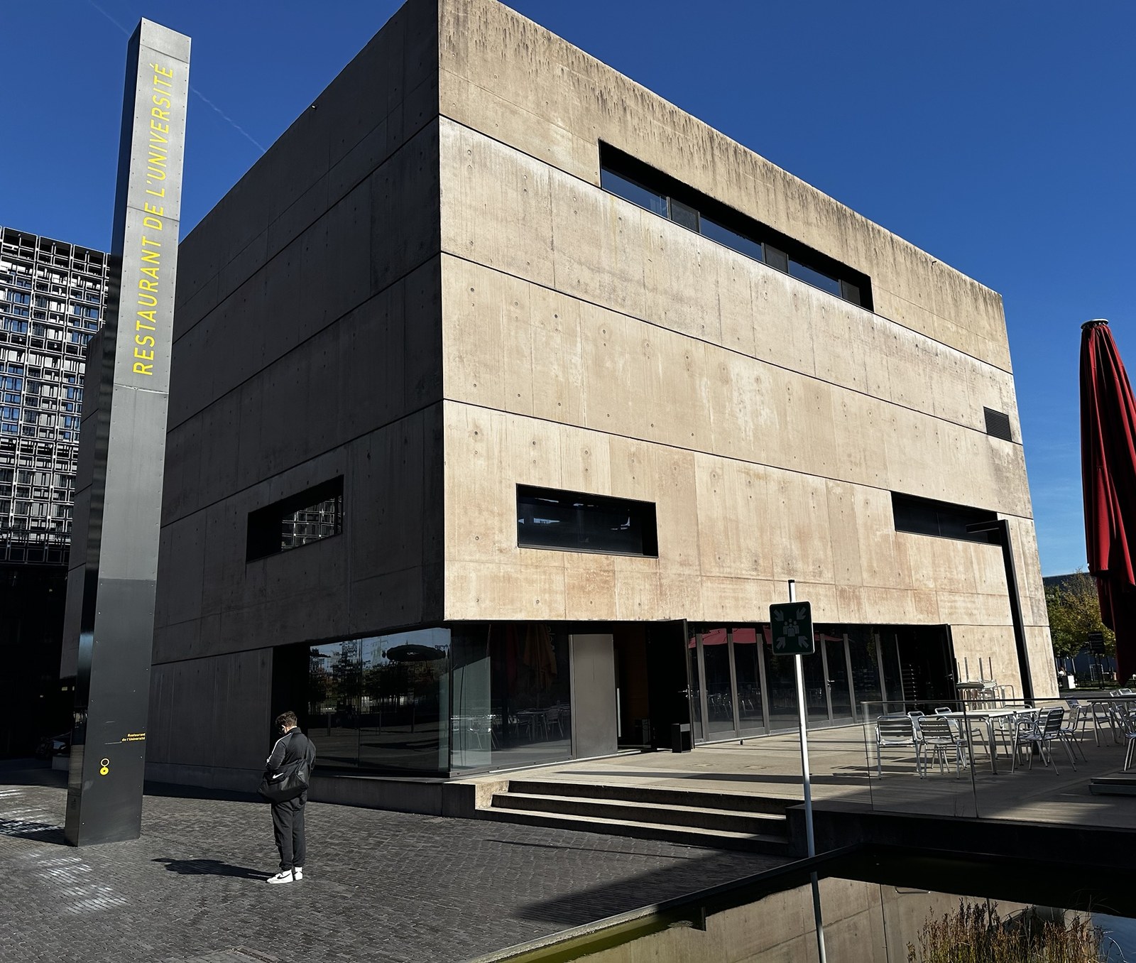 The Restaurant de l’Université building on the Belval campus, a concrete block with its name on a tall totem and a terrace of tables under red parasols beside the water.