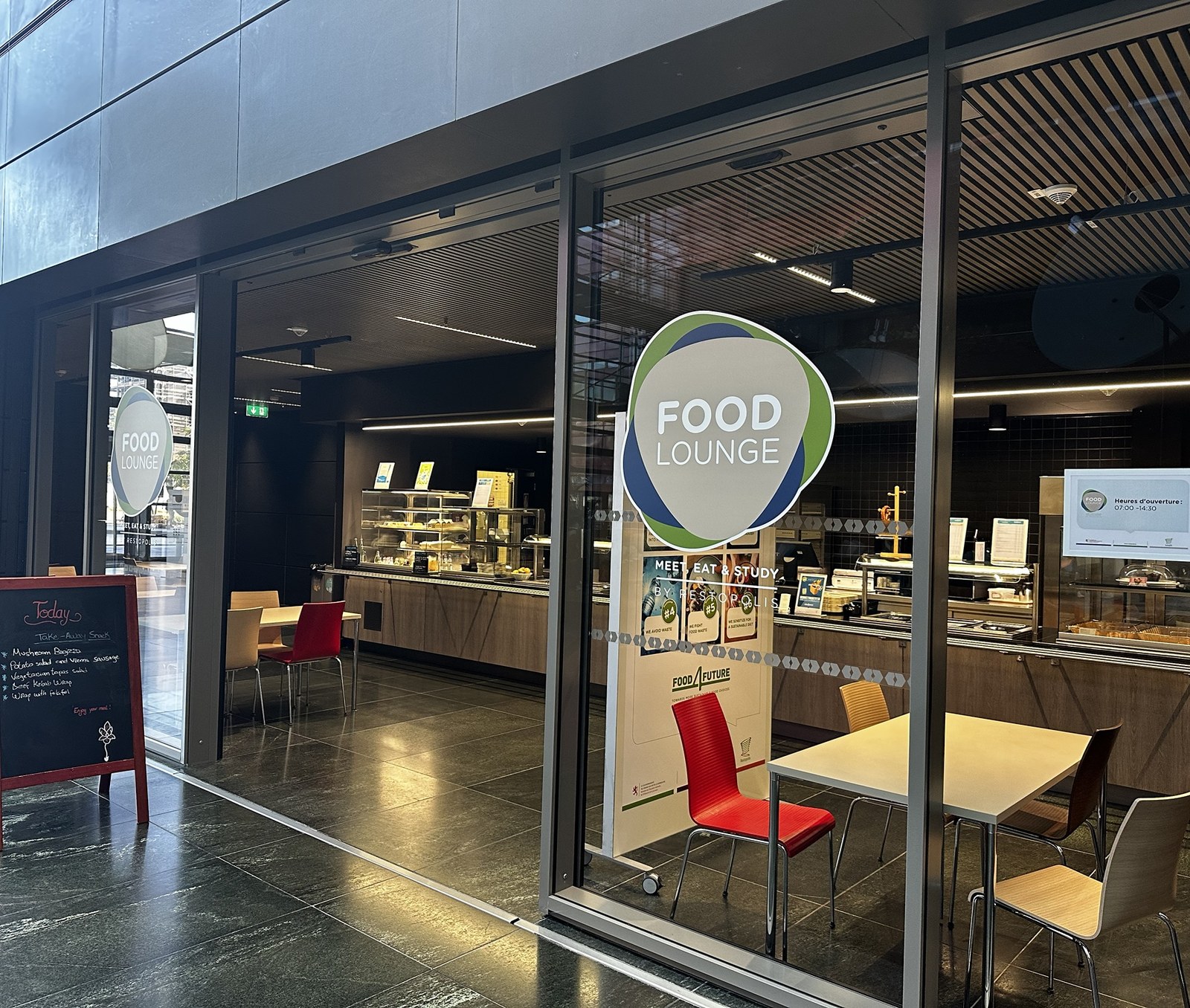 The Food Lounge counter in the Terres Rouges building, the green and blue Food Lounge roundel on the glass with MEET, EAT & STUDY by Restopolis beneath it, and the servery behind.