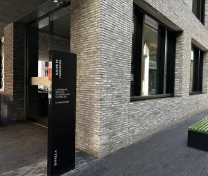 The black totem at Entrée A of the Maison de l’Innovation on Avenue des Hauts-Fourneaux in Belval, naming the Luxembourg Institute of Science and Technology and Luxinnovation, with the pale brick facade and green benches beside it.