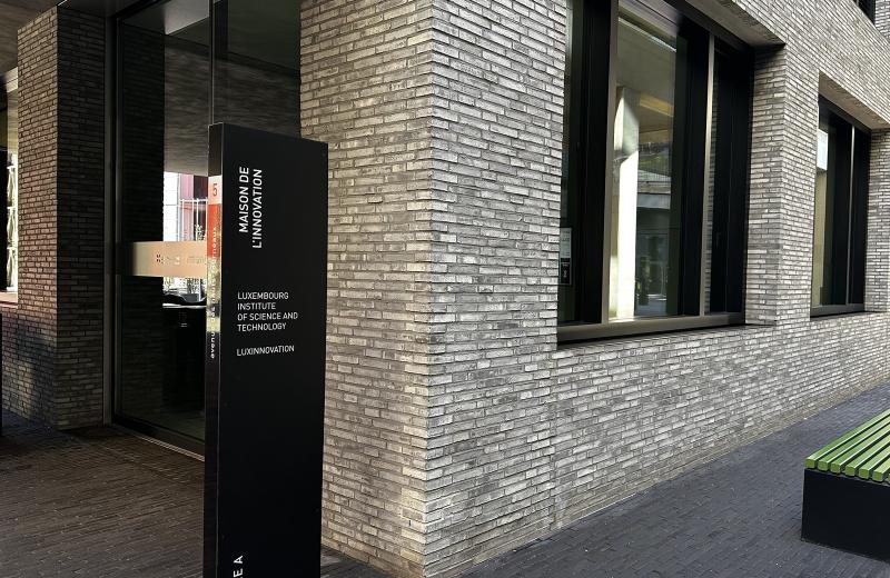 The black totem at Entrée A of the Maison de l’Innovation on Avenue des Hauts-Fourneaux in Belval, naming the Luxembourg Institute of Science and Technology and Luxinnovation, with the pale brick facade and green benches beside it.