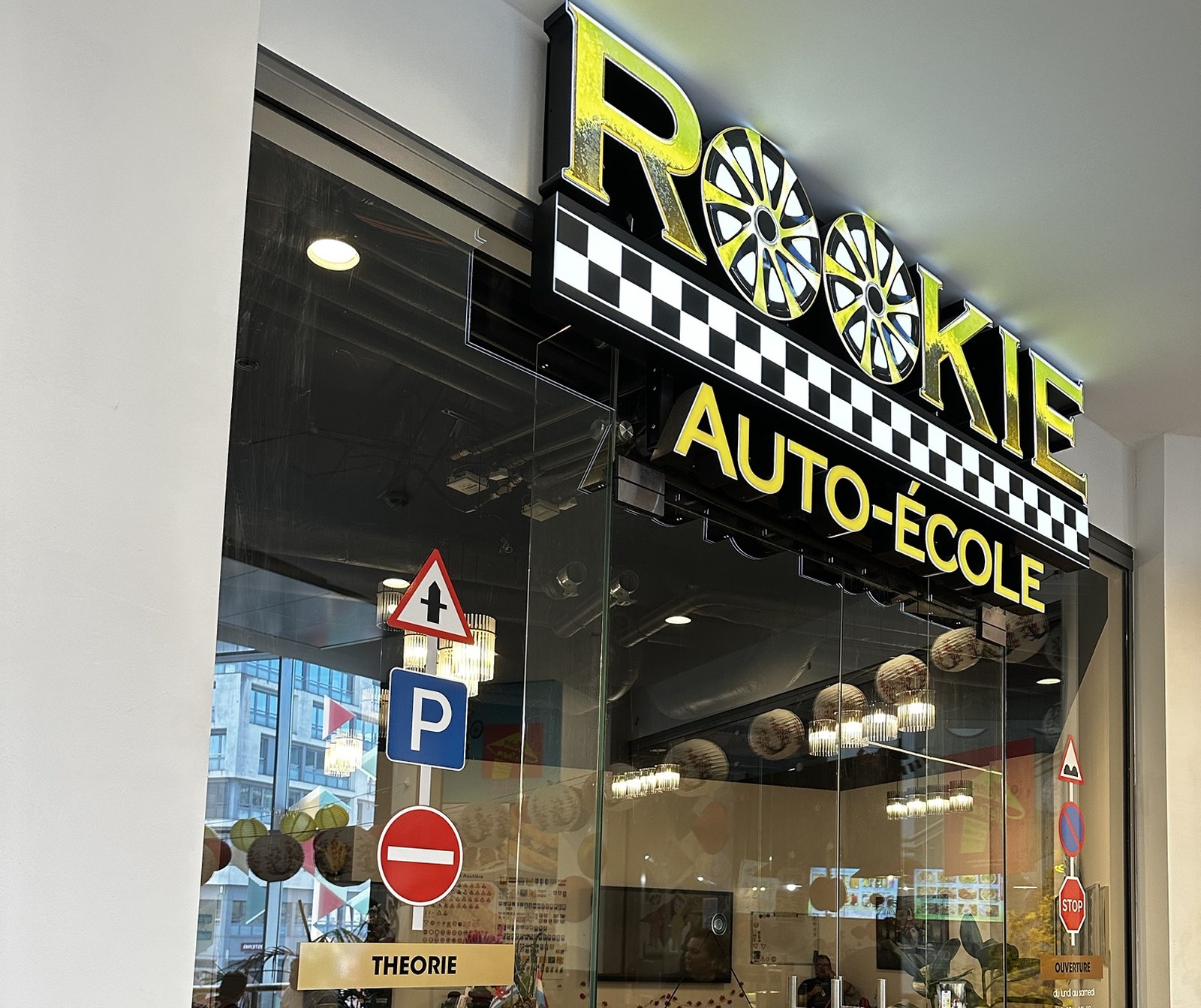 The Rookie Auto-École shopfront in Belval Plaza II, a yellow and black fascia with two wheel trims in place of the double O, road signs in the window and a classroom of yellow chairs inside.
