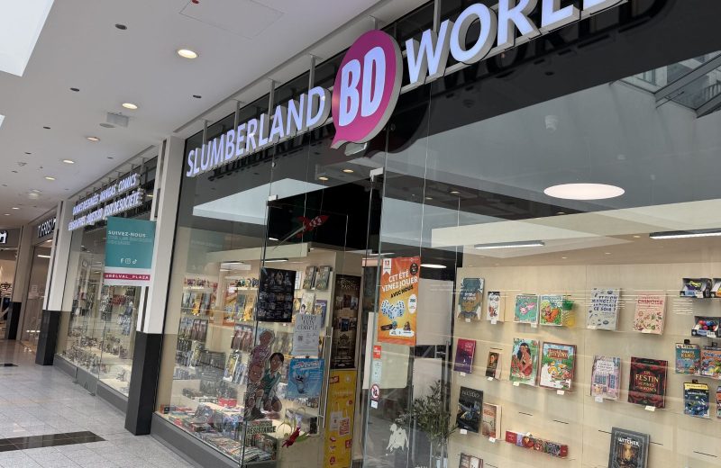 Slumberland BD World shopfront at Belval Plaza, a black and pink storefront with "Slumberland BD World" lettering above a shelf display of comics, manga, board games and collectible figures, a poster advertising a summer games event, and a teal sticker inviting shoppers to follow the centre's own social media accounts.