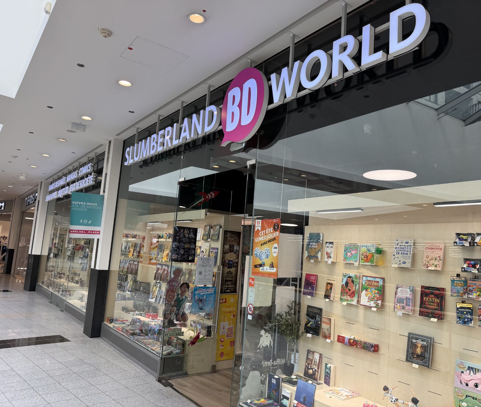 Slumberland BD World shopfront at Belval Plaza, a black and pink storefront with "Slumberland BD World" lettering above a shelf display of comics, manga, board games and collectible figures, a poster advertising a summer games event, and a teal sticker inviting shoppers to follow the centre's own social media accounts.