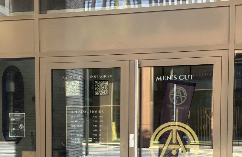 The Artists Barber salon doors, with a gold circular emblem and "Men's Cut" and "The Artists" lettering, opening hours listed in gold text for Monday to Saturday with Sunday marked closed, and QR codes for Salonkee booking and Instagram, with an interior poster listing haircut prices including a students category visible through the glass.
