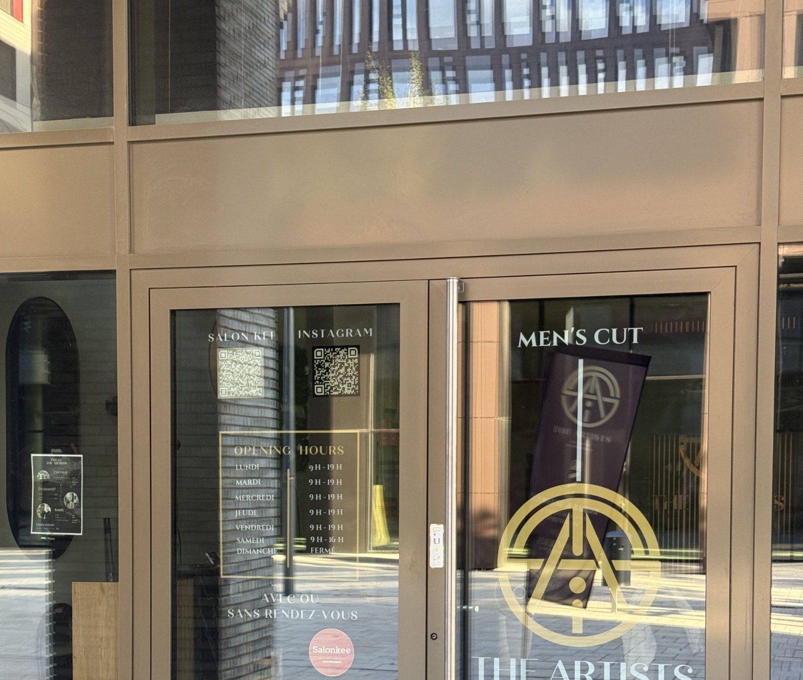 The Artists Barber salon doors, with a gold circular emblem and "Men's Cut" and "The Artists" lettering, opening hours listed in gold text for Monday to Saturday with Sunday marked closed, and QR codes for Salonkee booking and Instagram, with an interior poster listing haircut prices including a students category visible through the glass.